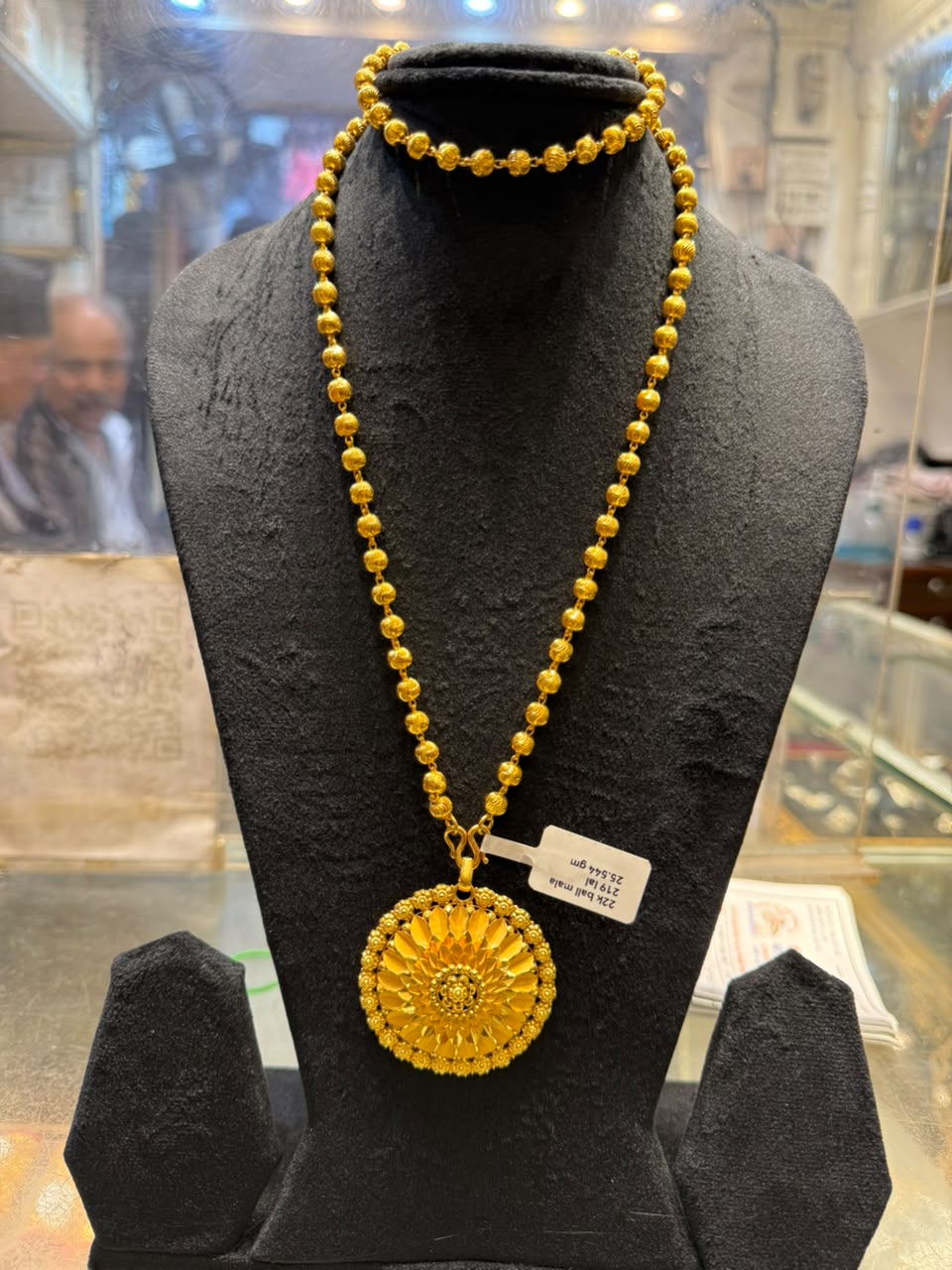 Gold ball mala with a round sunburst pendant; a shop tag is tied to the chain